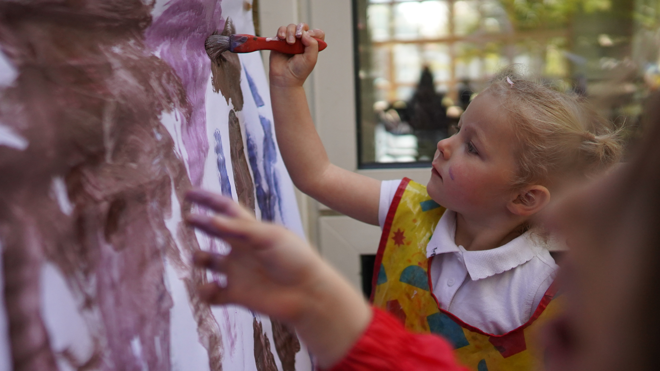 A young girl can be seen painting on a large sheet of paper on the wall with a paintbrush, whilst wearing an art apron.